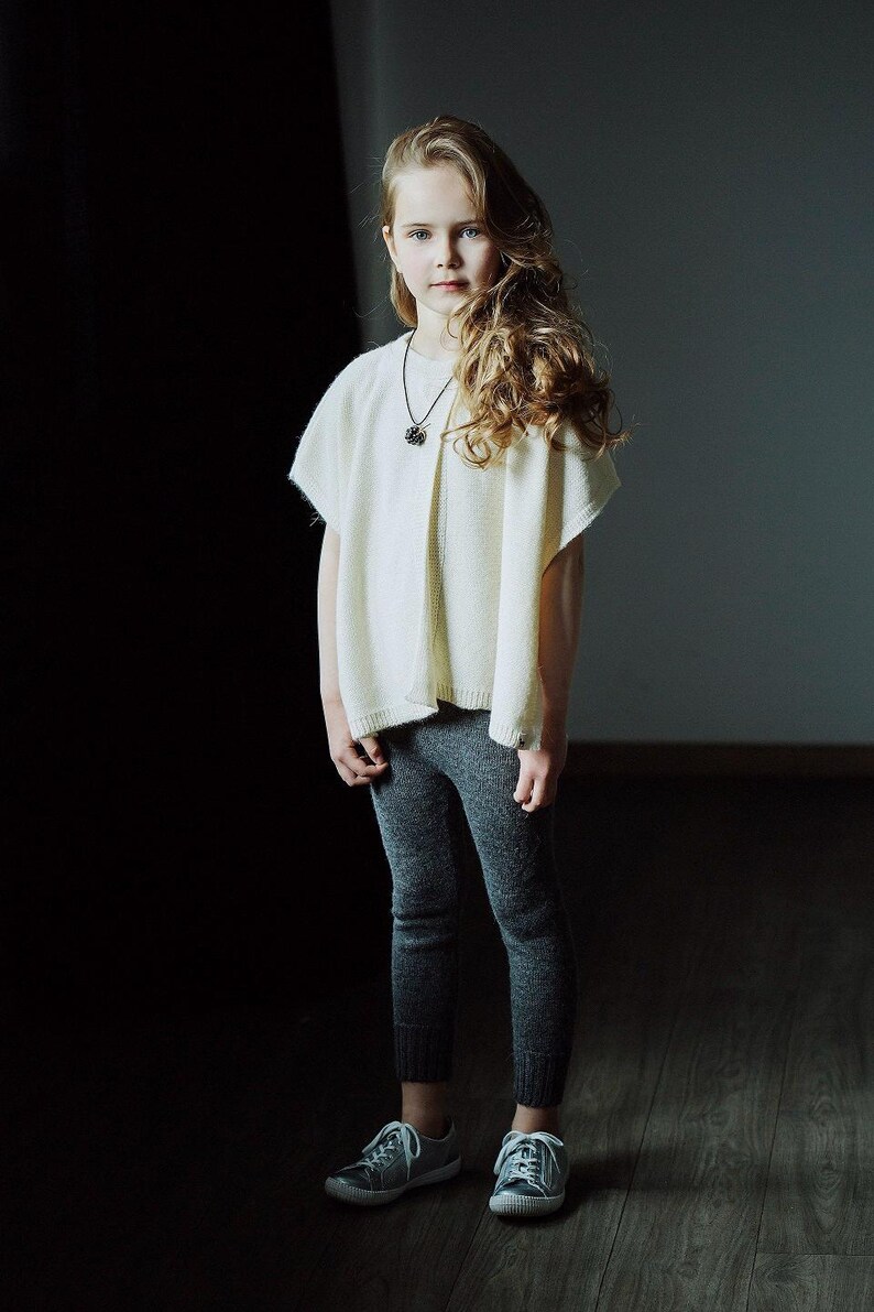 May include: A young girl with long blonde hair wearing a white sweater and grey pants. She is standing in front of a dark background and looking at the camera.