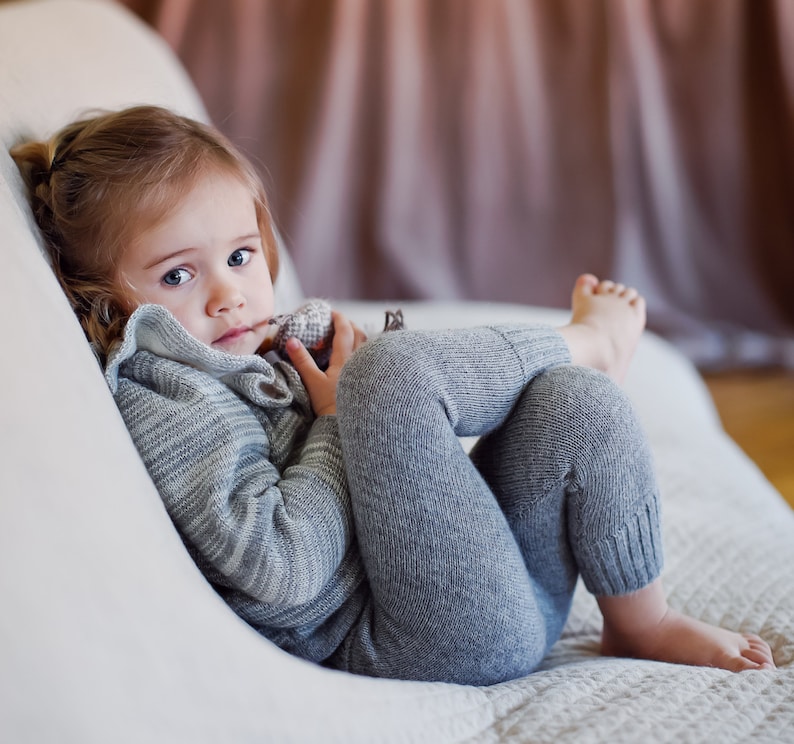 May include: A young child wearing a gray and white striped sweater and gray pants sits on a white couch. The child is holding a small stuffed animal in their hands.
