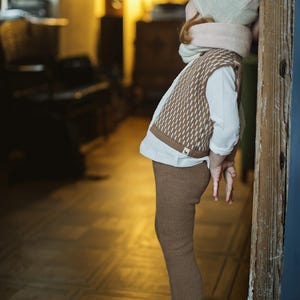 May include: A young child wearing a brown and white knitted vest, brown knitted pants, a white shirt, and a light gray knitted hat with a pink scarf. The child is standing near a wooden door.
