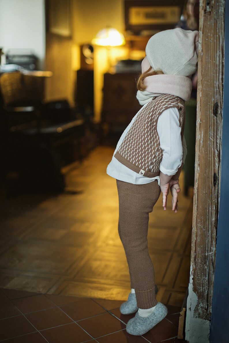 May include: A young child wearing a brown and white knitted vest, brown knitted pants, a white shirt, and a light gray knitted hat with a pink scarf. The child is standing near a wooden door.