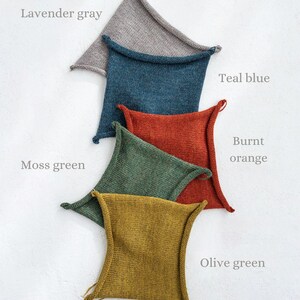 May include: Five knitted squares in different colors, stacked on top of each other. The colors are lavender gray, teal blue, burnt orange, moss green, and olive green.