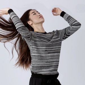 May include: A woman with long brown hair wearing a black and white striped sweater. She is standing with her arms raised and her hair is flowing behind her.
