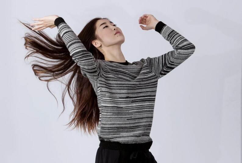 May include: A woman with long brown hair wearing a black and white striped sweater. She is standing with her arms raised and her hair is flowing behind her.