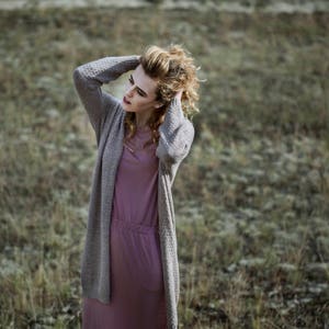 Puede incluir: Una mujer con el pelo largo, ondulado y casta&ntilde;o claro lleva un c&aacute;rdigan largo gris sobre un vestido rosa. Est&aacute; de pie en un campo de hierba verde.
