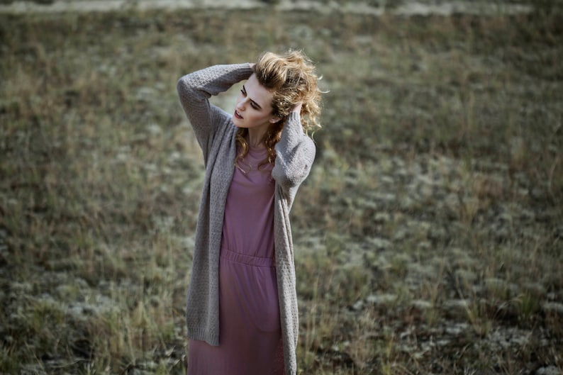 Puede incluir: Una mujer con el pelo largo, ondulado y casta&ntilde;o claro lleva un c&aacute;rdigan largo gris sobre un vestido rosa. Est&aacute; de pie en un campo de hierba verde.