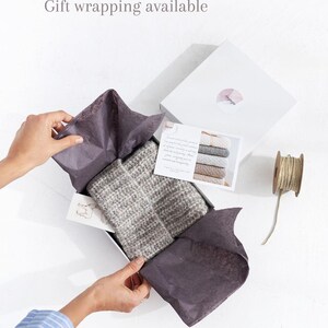 May include: A gray and white striped knitted scarf is folded and placed in a white gift box with gray tissue paper. The box is open and the tissue paper is being pulled back to reveal the scarf. A small card with text is visible in the box. The box is sitting on a white surface.