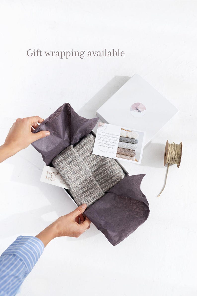 May include: A gray and white striped knitted scarf is folded and placed in a white gift box with gray tissue paper. The box is open and the tissue paper is being pulled back to reveal the scarf. A small card with text is visible in the box. The box is sitting on a white surface.