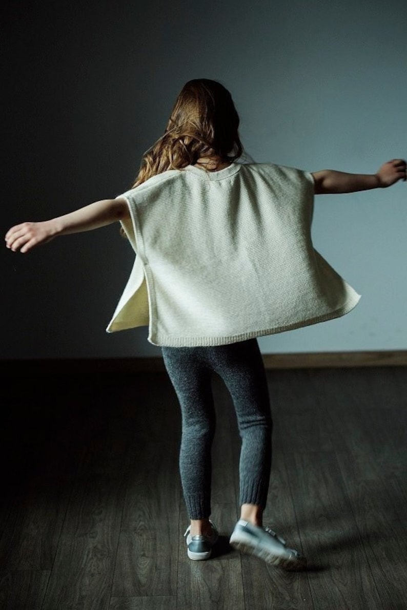 May include: A young person wearing a white knitted poncho with long sleeves and a grey pair of leggings. They are standing on a wooden floor with their arms outstretched.