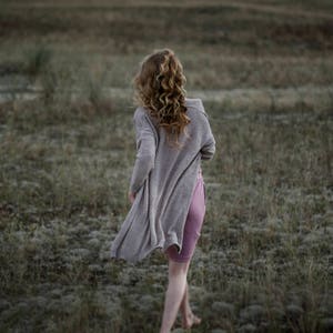 Puede incluir: Una mujer con el pelo largo y rubio rizado camina de espaldas a la c&aacute;mara en un campo. Lleva un c&aacute;rdigan gris claro y una falda rosa.