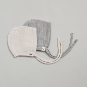 May include: Two baby bonnets, one white and one gray, with long ties. Both bonnets have a small llama logo.