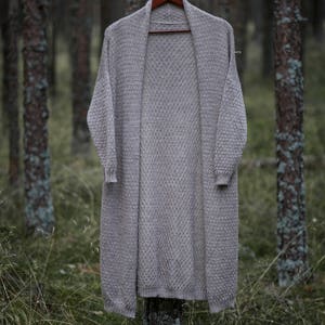 Puede incluir: Un c&aacute;rdigan largo de color gris claro colgado de una percha de madera en un entorno de bosque. El c&aacute;rdigan tiene un corte holgado y un dise&ntilde;o sencillo.