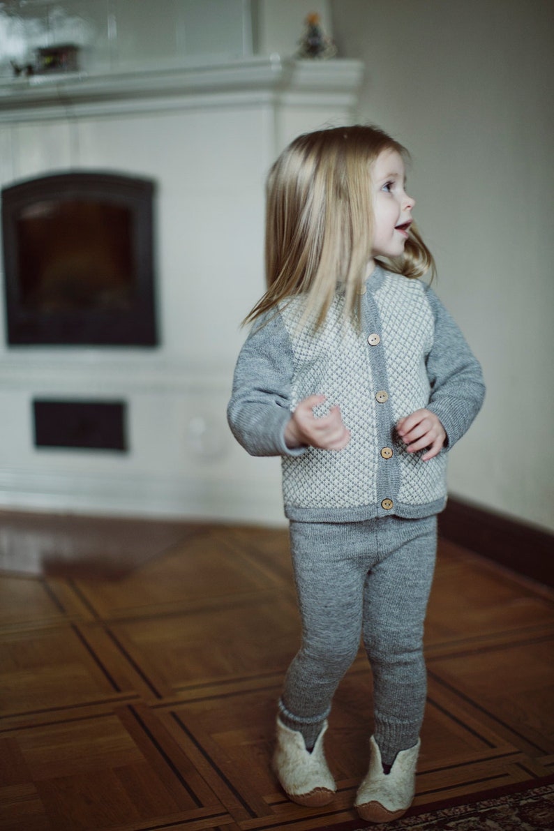 May include: A young girl wearing a gray and white knitted sweater with buttons and matching gray knitted pants. She is standing on a wooden floor and looking to the right.