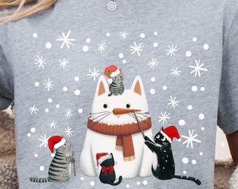 May include: A light gray t-shirt featuring a whimsical Christmas design. The graphic depicts a snowman cat with a brown scarf, surrounded by other cats wearing Santa hats and snowflakes. A mug with a Santa Claus illustration is also visible.