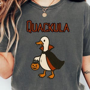 Könnte beinhalten: Dunkelgraues T-Shirt mit dem Wort "Quackula" in orangefarbenen und braunen Buchstaben. Auf der Vorderseite ist eine Cartoon-Ente mit einem Vampirumhang und einem Kürbis-Eimer abgebildet. Ein lustiges Halloween-Grafik-T-Shirt.