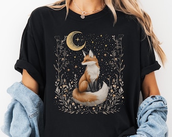 May include: Black t-shirt featuring a watercolor illustration of a fox with orange and white fur, sitting beneath a gold crescent moon and stars. The design is surrounded by floral elements in shades of gold and gray.
