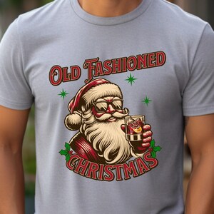 K&ouml;nnte beinhalten: Hellgraues T-Shirt mit einer Grafik von Santa Claus mit Sonnenbrille und einem Cocktail. Der Text "Old Fashioned Christmas" ist in Rot und Wei&szlig;, mit gr&uuml;nen Sternakzenten. Das Design ist festlich und weihnachtlich.