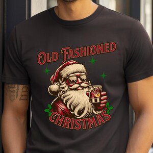 K&ouml;nnte beinhalten: Schwarzes T-Shirt mit einer Illustration von Santa Claus, der eine Sonnenbrille tr&auml;gt und einen Cocktail h&auml;lt. Der Text "Old Fashioned Christmas" ist oberhalb und unterhalb der Santa-Grafik angeordnet. Gr&uuml;ne Sternakzente sind enthalten.