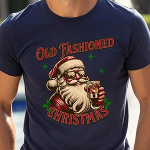 K&ouml;nnte beinhalten: Marineblaues T-Shirt mit einer Grafik von Santa Claus mit Sonnenbrille und einem Cocktail. Der Text "Old Fashioned Christmas" ist in Rot und Gr&uuml;n &uuml;ber der Santa-Illustration abgebildet.