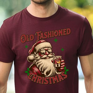 K&ouml;nnte beinhalten: Burgunderfarbenes T-Shirt mit einer Grafik von Santa Claus mit Sonnenbrille und einem Cocktail. Der Text "Old Fashioned Christmas" ist in Rot und Gr&uuml;n &uuml;ber und unter der Santa-Illustration angeordnet.