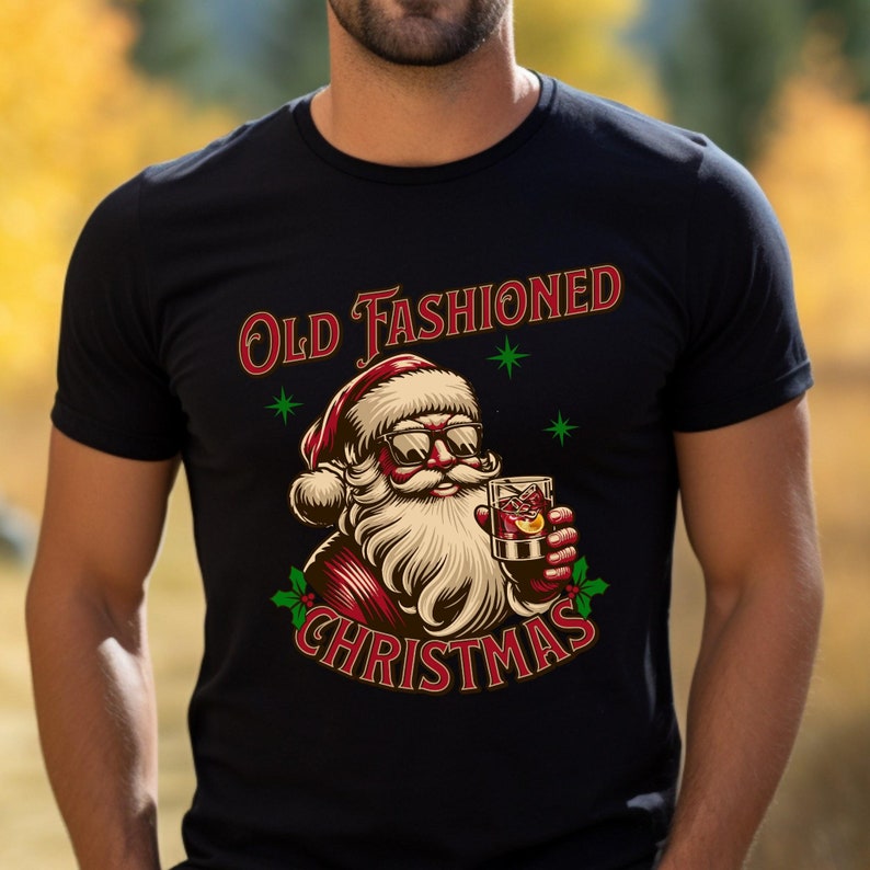 K&ouml;nnte beinhalten: Schwarzes T-Shirt mit einer Grafik von Santa Claus mit Sonnenbrille und einem Cocktail. Der Text "Old Fashioned Christmas" ist in Rot und Gr&uuml;n &uuml;ber und unter der Santa-Illustration abgebildet.