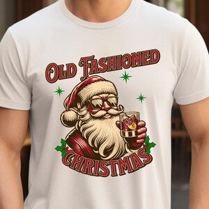 K&ouml;nnte beinhalten: Wei&szlig;es T-Shirt mit einem Cartoon-Weihnachtsmann, der eine Sonnenbrille tr&auml;gt und einen Cocktail h&auml;lt. Der Aufdruck "Old Fashioned Christmas" ist rot und gr&uuml;n, mit gr&uuml;nen Sternen.