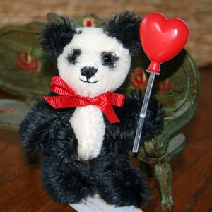 Puede incluir: Un pequeño peluche de oso panda con pelaje blanco y negro, un lazo rojo y un globo rojo en forma de corazón. El panda sostiene el globo en un palo transparente. El fondo incluye una silla verde.