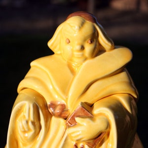 May include: A yellow ceramic figurine of a monk holding a book. The monk is wearing a brown hood and robe.