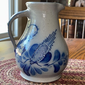 May include: A light grey stoneware pitcher with a speckled finish and a blue hand-painted floral design. The pitcher features a curved handle, a wide base, and a pouring spout. The blue pattern includes intricate petal shapes and a textured centre.
