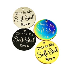 May include: Four round metal tags with the text "This is My Soft Girl Era" engraved on them. The tags are in different colors: silver, rainbow, black, and gold.
