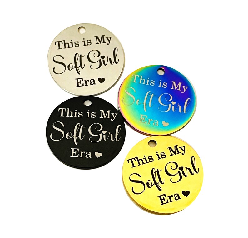 May include: Four round metal tags with the text "This is My Soft Girl Era" engraved on them. The tags are in different colors: silver, rainbow, black, and gold.