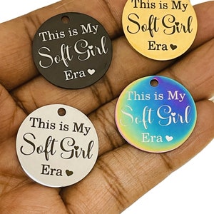 May include: Four round metal tags with the text "This is My Soft Girl Era" engraved on them. The tags are in different colors: black, gold, silver, and iridescent.