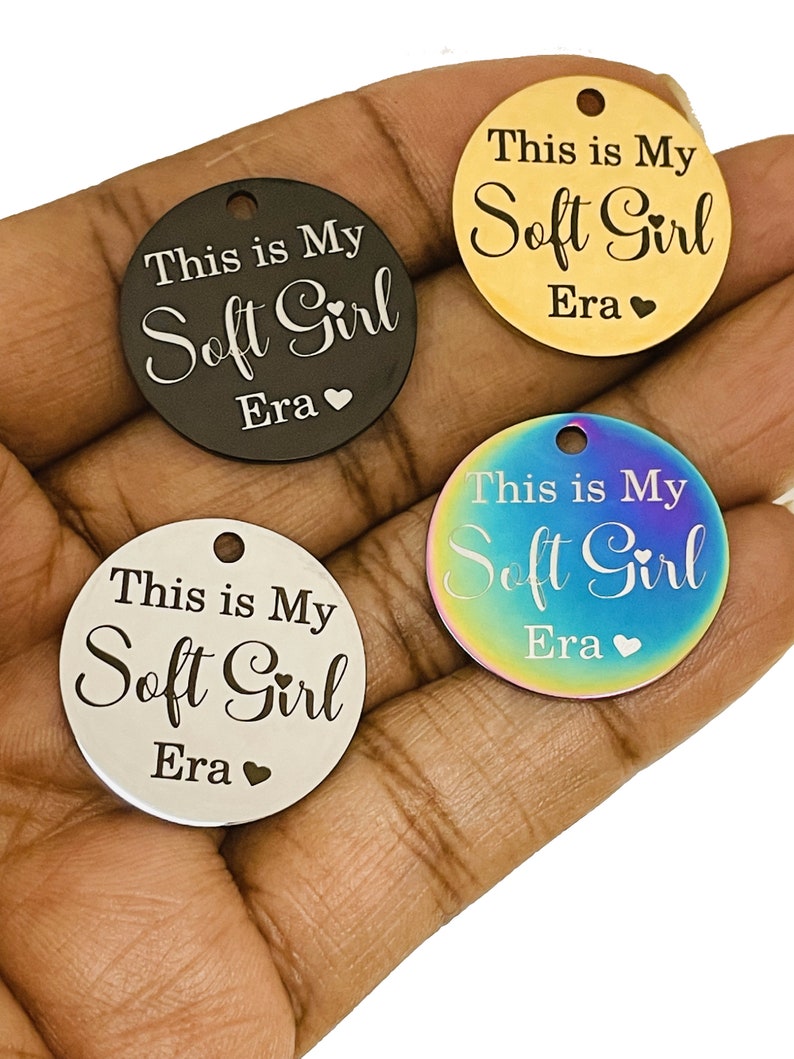 May include: Four round metal tags with the text "This is My Soft Girl Era" engraved on them. The tags are in different colors: black, gold, silver, and iridescent.