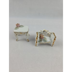May include: Miniature white porcelain furniture set, including a small table and a loveseat. The table and loveseat have gold trim and are decorated with pink roses and green leaves. The set is designed for a dollhouse or miniature display.