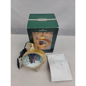May include: A gold-toned Hallmark Keepsake ornament with a recessed circular scene featuring two figures and a dog in a snowy landscape. The ornament includes a black power cord, its original green box with product imagery, and a white instruction manual.