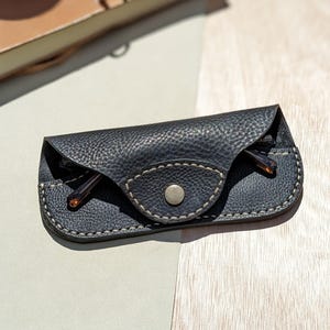 May include: A black leather glasses case with a snap closure and a stitched design. The case is closed and a pair of glasses is inside.