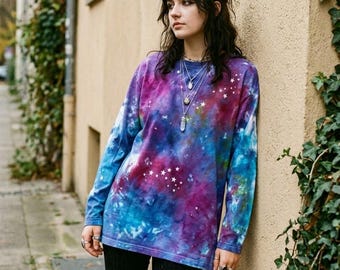 May include: A long-sleeve crew neck shirt featuring a tie-dye pattern in shades of deep purple, magenta, and bright blue. The fabric is decorated with small, white, printed star constellations scattered across the front and sleeves.