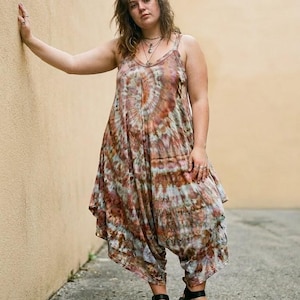 Op de afbeelding: Een mouwloze jumpsuit die tot de enkels valt, voorzien van een tie-dye patroon in bruine, roestkleurige en gedempte grijstinten. Het kledingstuk heeft een ronde hals, dunne schouderbandjes en een losvallend model met een laag kruis.