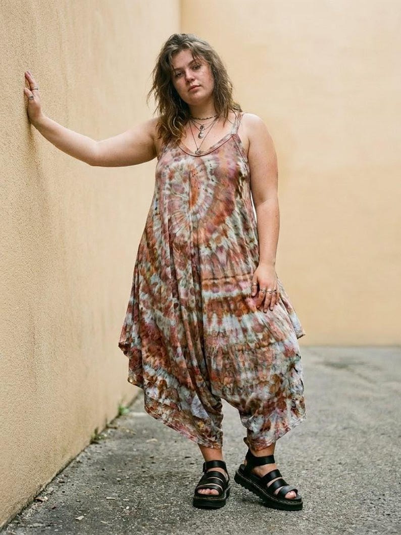 Op de afbeelding: Een mouwloze jumpsuit die tot de enkels valt, voorzien van een tie-dye patroon in bruine, roestkleurige en gedempte grijstinten. Het kledingstuk heeft een ronde hals, dunne schouderbandjes en een losvallend model met een laag kruis.