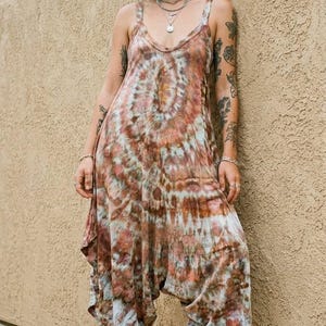 Op de afbeelding: Een mouwloze jumpsuit met een tie-dye patroon in bruin-, roest- en cr&egrave;metinten. Het kledingstuk heeft een ronde halslijn, brede schouderbanden en een losvallend model met een verlaagd kruis dat taps toeloopt naar de enkels, gecombineerd met zwarte sandalen.