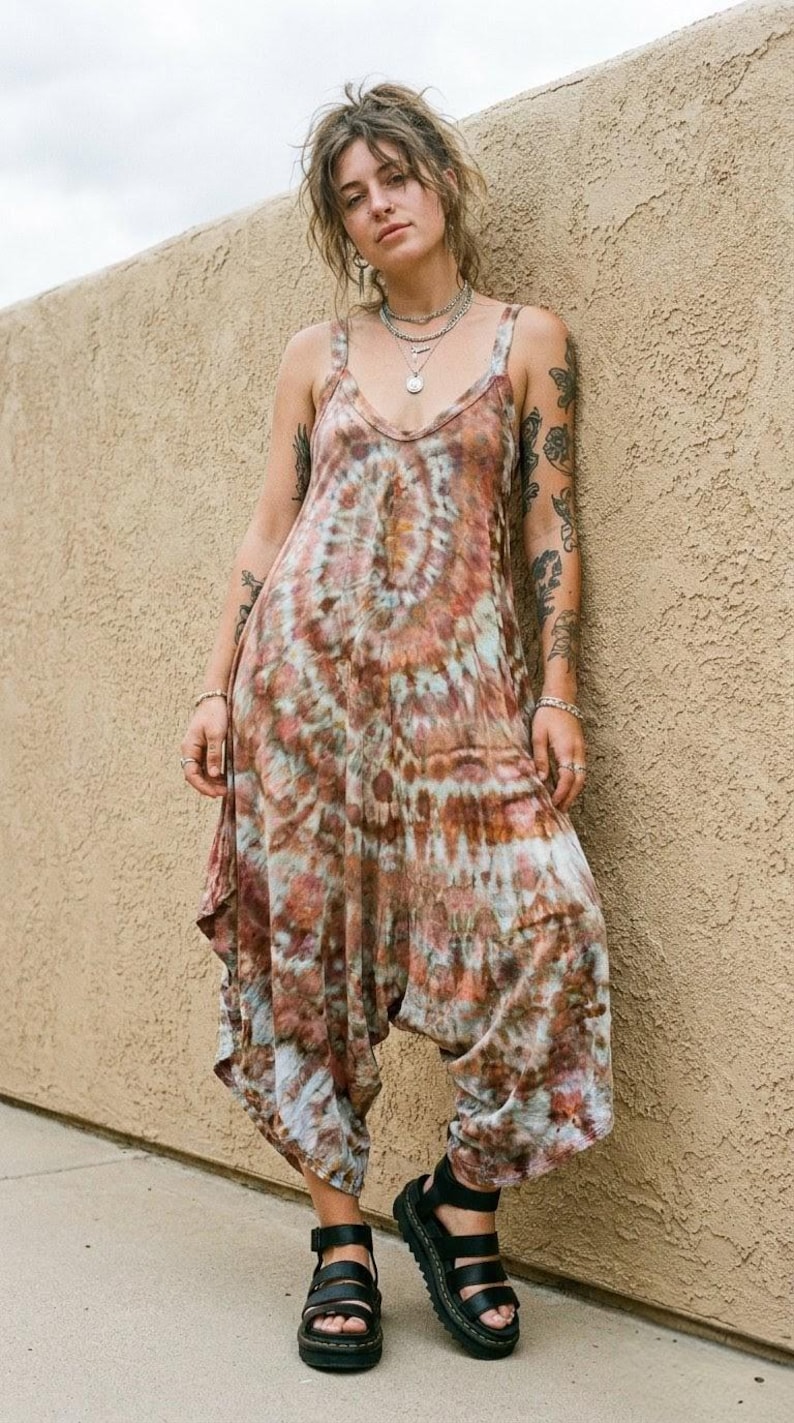 Op de afbeelding: Een mouwloze jumpsuit met een tie-dye patroon in bruin-, roest- en cr&egrave;metinten. Het kledingstuk heeft een ronde halslijn, brede schouderbanden en een losvallend model met een verlaagd kruis dat taps toeloopt naar de enkels, gecombineerd met zwarte sandalen.