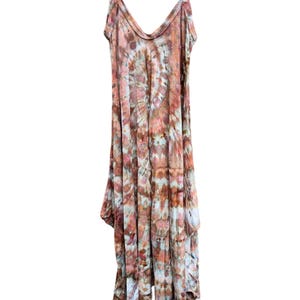Op de afbeelding: Een mouwloze jurk met een tie-dye patroon in tinten roest, bruin, cr&egrave;me en gedempt blauw. Het kledingstuk heeft een diepe V-hals, dunne schouderbandjes en een asymmetrische zoom die op verschillende lengtes rond de onderkant valt.