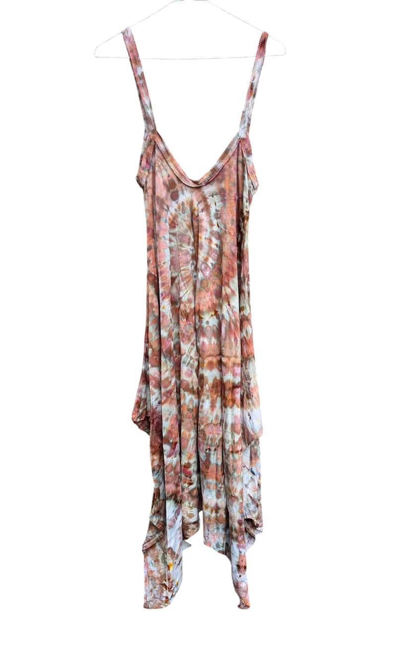 Op de afbeelding: Een mouwloze jurk met een tie-dye patroon in tinten roest, bruin, cr&egrave;me en gedempt blauw. Het kledingstuk heeft een diepe V-hals, dunne schouderbandjes en een asymmetrische zoom die op verschillende lengtes rond de onderkant valt.