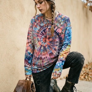 Peut inclure: Un sweat-shirt à manches longues et col rond arborant un motif tie-dye en spirale aux tons bleu, rose, orange et jaune. Le vêtement est associé à un pantalon en velours côtelé noir usé, un sac à dos en cuir marron et un bonnet tie-dye.