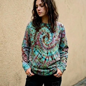 Op de afbeelding: Een shirt met lange mouwen en een spiraalvormig tie-dye patroon in tinten blauwgroen, bruin en gedempt paars. Het shirt heeft een ronde hals en lange mouwen, gecombineerd met een zwarte spijkerbroek met versleten details bij de zakken en knieën.
