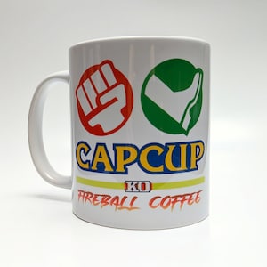 May include: White ceramic coffee mug with a red fist, green check mark, and blue and yellow "CAPCUP KO FIREBALL COFFEE" text.