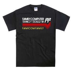 Op de afbeelding: Zwart T-shirt met een wit en rood grafisch ontwerp met de tekst "FAMILY COMPUTER" en "ファミリ―コンピュータ" in het Japans. De grafiek bevat ook een rood en wit pijlontwerp en de tekst "FAMICOM FAMILY" in geel.