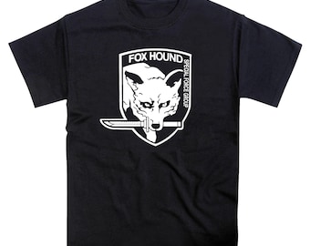 May include: A black short-sleeve t-shirt featuring a white graphic print of a fox head holding a knife in its mouth inside a shield emblem. The text FOX HOUND and SPECIAL FORCE GROUP is printed in white within the shield design on the chest.