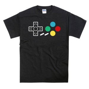 Op de afbeelding: Zwart T-shirt met een witte afbeelding van een videogamecontroller met vier gekleurde knoppen. De knoppen zijn rood, geel, groen en blauw.