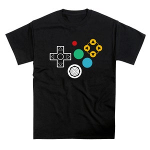 May include: Black t-shirt with a white gamepad design featuring a directional pad, four colored buttons, and a circular button. The buttons are red, green, blue, and yellow.