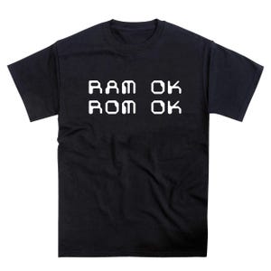 May include: Black crew neck t-shirt with white text. The text reads "RAM OK" on the top line and "ROM OK" on the bottom line. The shirt is made of a soft material.